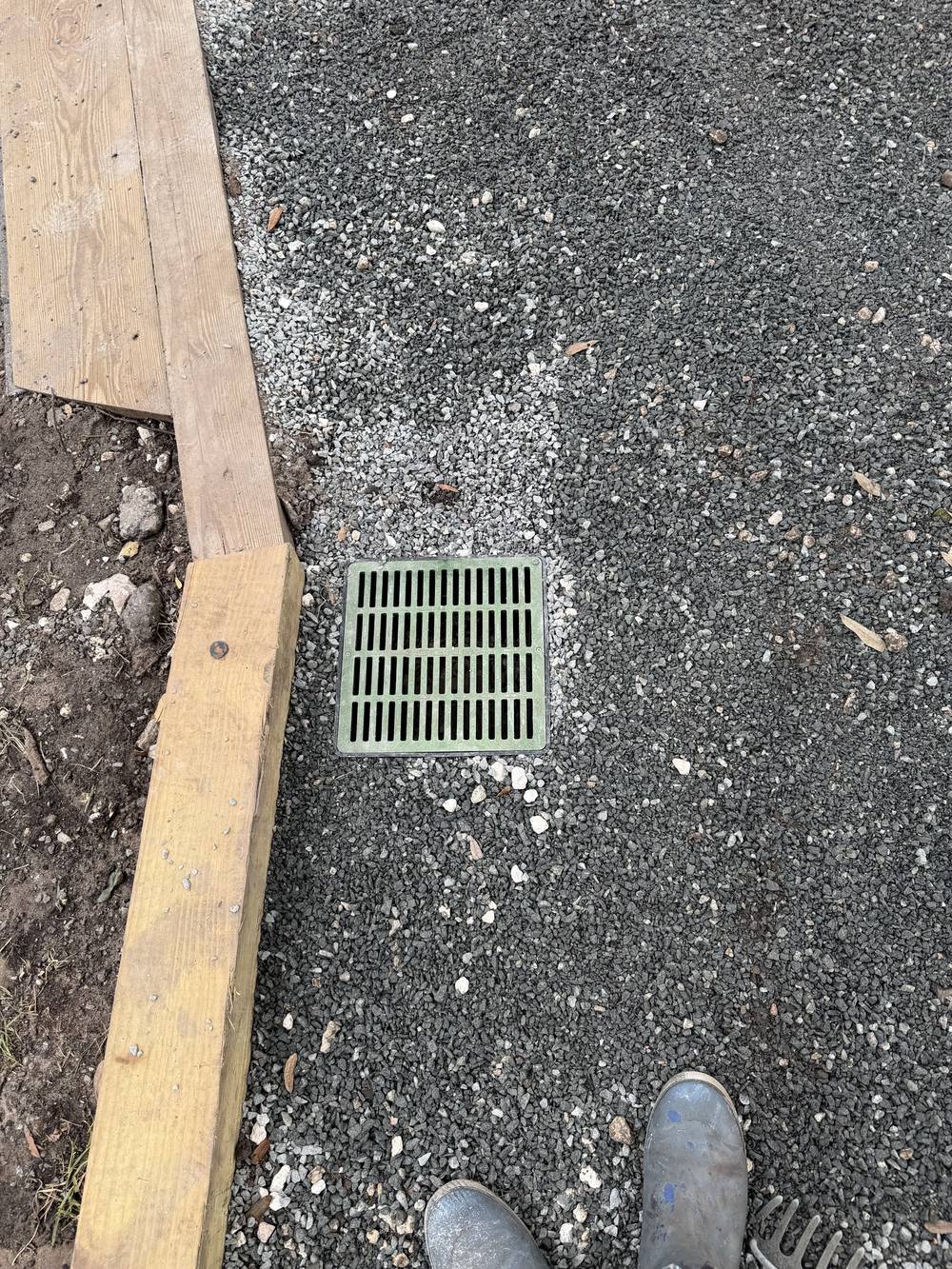 Square green catch basin grate set flush in a gravel yard beside timber edging