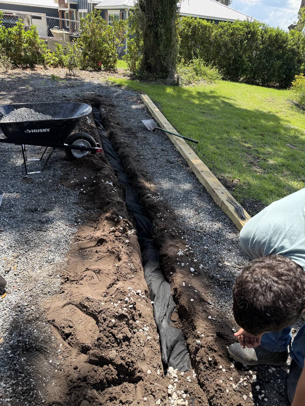Fabric-lined French drain trench dug through a gravel side yard, before the pipe and stone go in